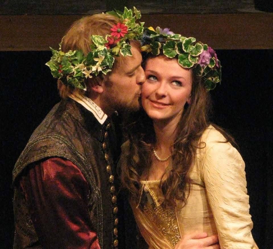 Rainbow on Mars co-director/performer Nate Bitton kisses creator/performer Devon Healey on the cheek; both are in period costume in this production still from their early years in theatre school at UTM Sheridan. Healey, crowned with a flower laurel on top of long, curled brown hair, smiles and looks off right coyly, while Bitton, also wearing a flower laurel over sandy blonde hair, kisses Healey's cheek, eyes closed.